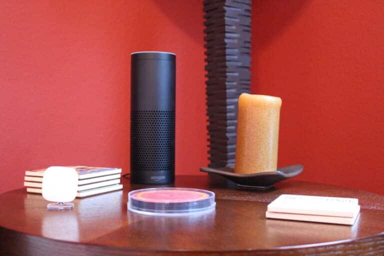A round wooden table with an Amazon Echo speaker, a candle, coasters, and small decorative items against a red wall.