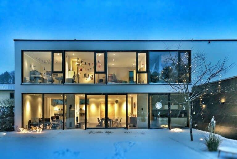 Modern two-story home with large windows glowing warmly, surrounded by snow.