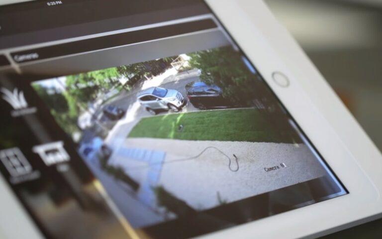 Smart home security camera feed displayed on tablet, showing driveway with vehicles and landscaped area, illustrating home automation and safety features.