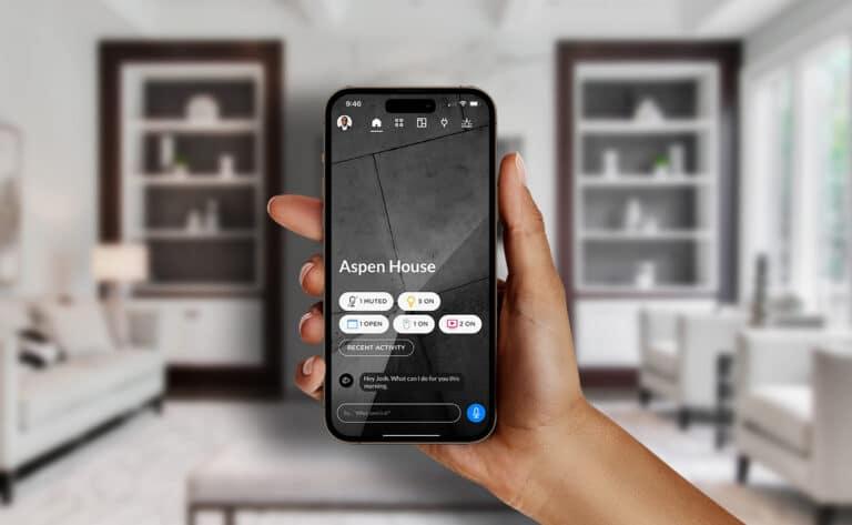 A hand holding a smartphone in a modern, elegant living room. The phone screen displays a smart home control app named "Aspen House," showing the status of devices such as lights, speakers, doors, and cameras. The interface includes voice command features