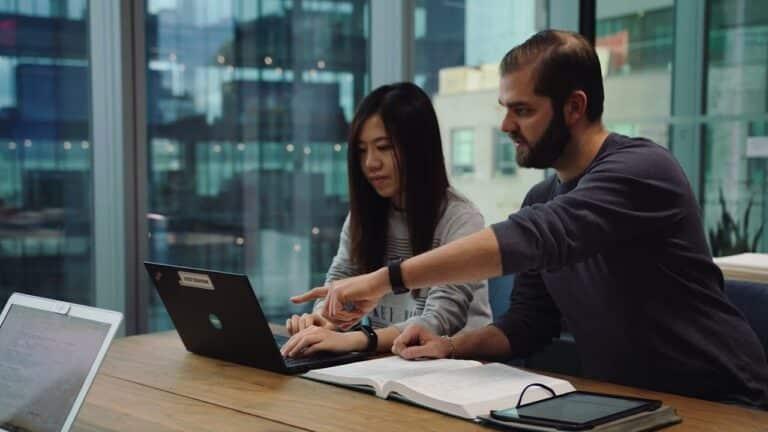 Two individuals collaborating at a wooden table, focused on a laptop, with a notebook and tablet nearby, illustrating remote work and technology integration in home settings.