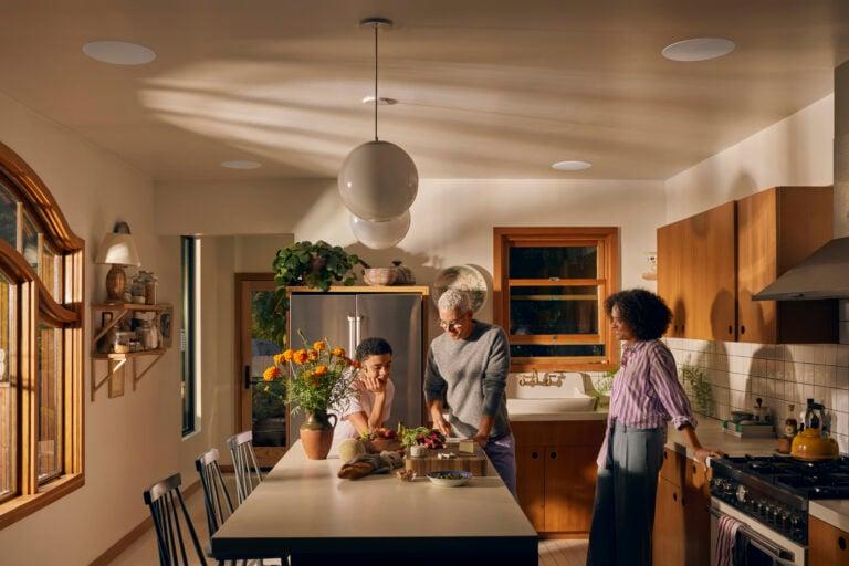 Three people enjoy time together in a warm, modern kitchen with natural wood accents. One person prepares food at the kitchen island while the others engage in conversation. The space features a vase of orange flowers, contemporary pendant lighting, woode