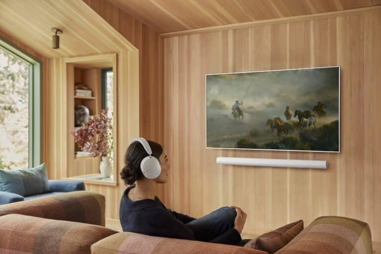 A woman wearing white over-ear headphones sits comfortably on a plaid-patterned couch in a cozy wooden-paneled living room, watching a wall-mounted TV displaying a painting of cowboys and horses. A white soundbar is mounted below the TV, and a window seat