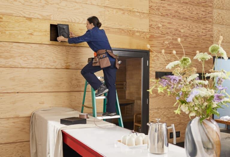 A woman on a ladder installing a speaker into a wooden wall inside a modern home, with tools around and flowers in the foreground.