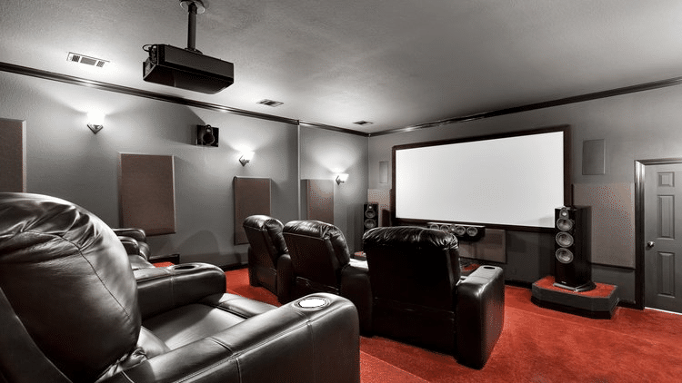 A luxurious home theater with leather recliners, a large projector screen, surround sound speakers, and a red carpeted floor.