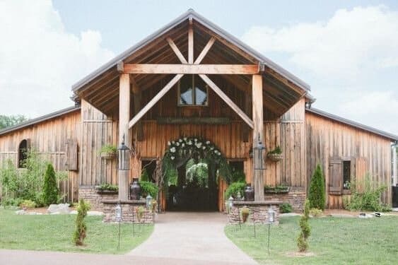 A rustic wooden barn-style venue with a large triangular entrance supported by wooden beams. The structure features natural wood siding, stone bases on the front pillars, and hanging plants. A paved walkway leads to the arched entryway, which is adorned with greenery and white floral decor, suggesting it is set up for an event like a wedding. The surrounding area is landscaped with green grass, shrubs, and small trees under a partly cloudy sky.