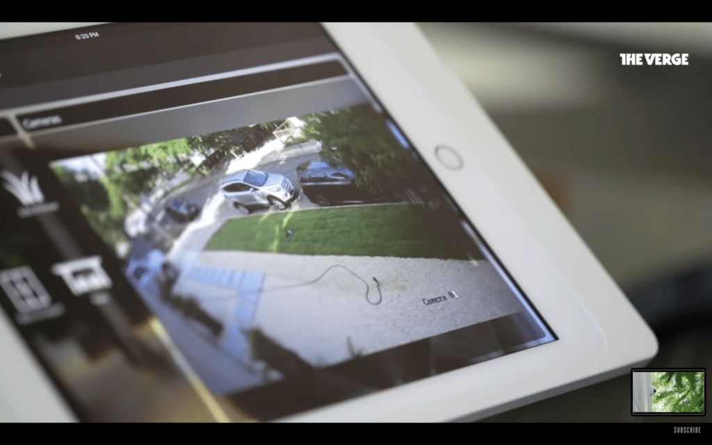 A close-up of a white tablet displaying a live security camera feed of a front yard with a driveway. The scene shows a few parked cars and a pathway with a garden hose laying across it. The interface appears to be part of a smart home system. The Verge logo is visible in the top-right corner of the screen.