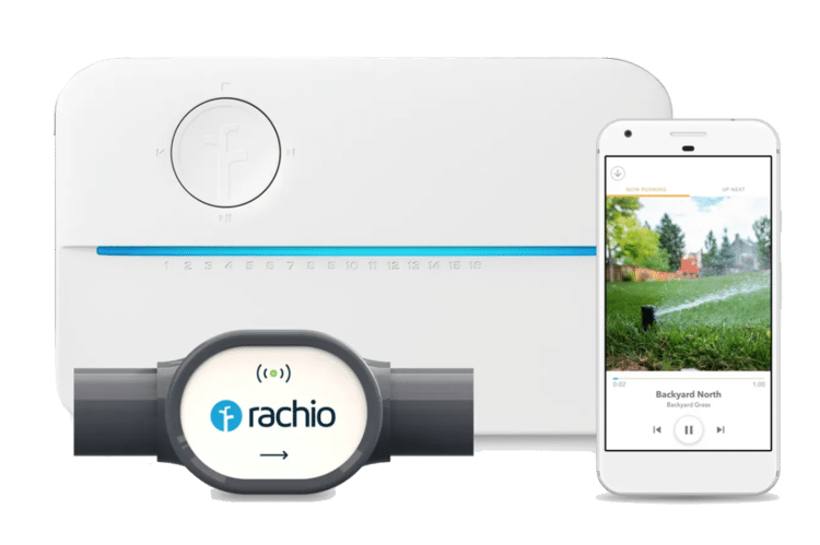 A Rachio smart irrigation system featuring three components: a white irrigation controller, a water flow meter labeled &ldquo;rachio,&rdquo; and a smartphone displaying the Rachio app interface. The app screen shows a sprinkler actively watering a backyard lawn with a status label reading "Backyard North.".