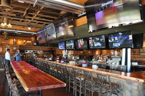 A rustic sports bar with multiple TVs, a long wooden bar, and high-top seating.