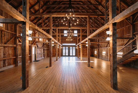 Interior of a spacious rustic barn featuring exposed wooden beams, high vaulted ceilings, and wide plank wood flooring. The space is illuminated by multiple wrought iron chandeliers and wall-mounted sconces, casting a warm, ambient glow. French doors with glass panels are located at the far end, allowing natural light to filter in. A wooden staircase on the right leads to an upper loft area, enhancing the barn's open and airy architectural design, ideal for events or gatherings.
