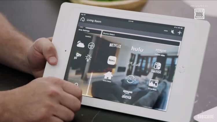 Person holding a tablet displaying a smart home interface with options to control entertainment sources like Netflix, Hulu, Amazon Video, Roku, PS4, Nintendo Switch, and smart home assistants like Alexa and Sonos.