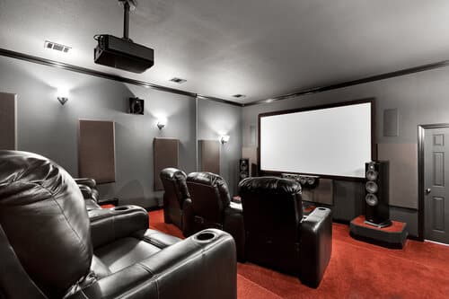 Modern home theater with black leather recliners, red carpet, acoustic panels on gray walls, large projection screen, and ceiling-mounted projector.