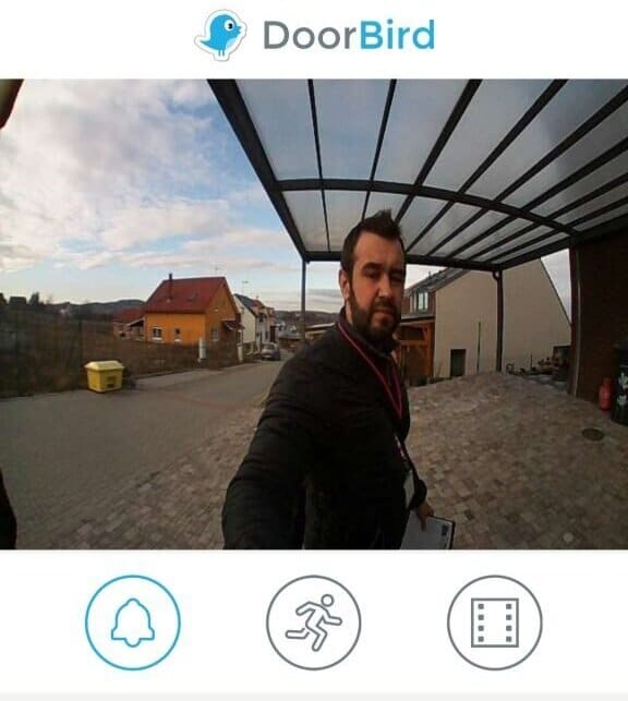Selfie of a man near a DoorBird video doorbell, showcasing smart home technology, with residential buildings in the background and interface icons for notifications, motion detection, and video recording.
