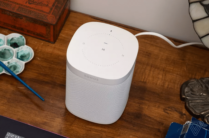 A white Sonos smart speaker placed on a wooden table with art supplies and decorative items nearby.
