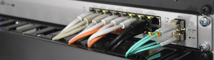 Close-up image of a network switch with multiple Ethernet and fiber optic cables connected. The cables are color-coded (white, orange, black, and teal), and the switch shows indicator lights above each port, signaling data activity and connectivity. The setup is mounted in a professional server rack, indicating a high-performance networking environment.