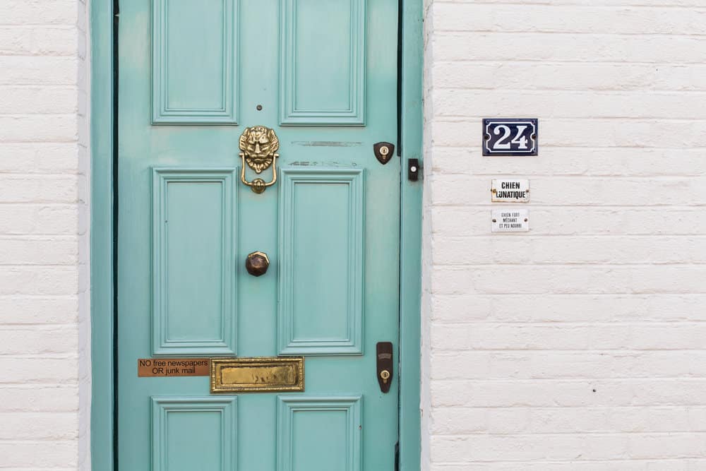 Turquoise front door with a brass knocker and mail slot, set in a white brick wall with house number 24.