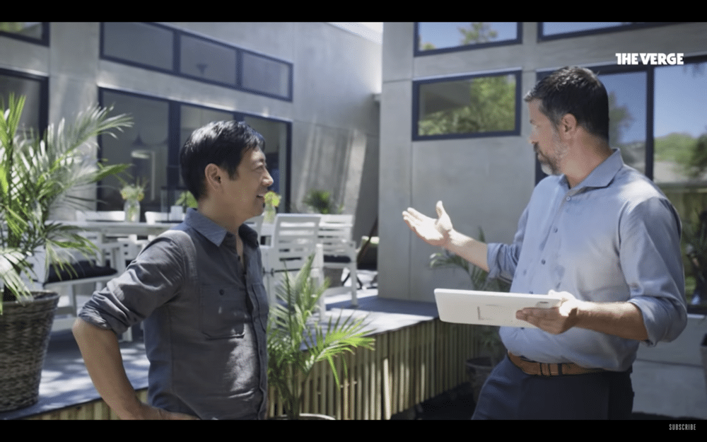 Two men having a discussion outside a modern home, with one holding a tablet and gesturing.