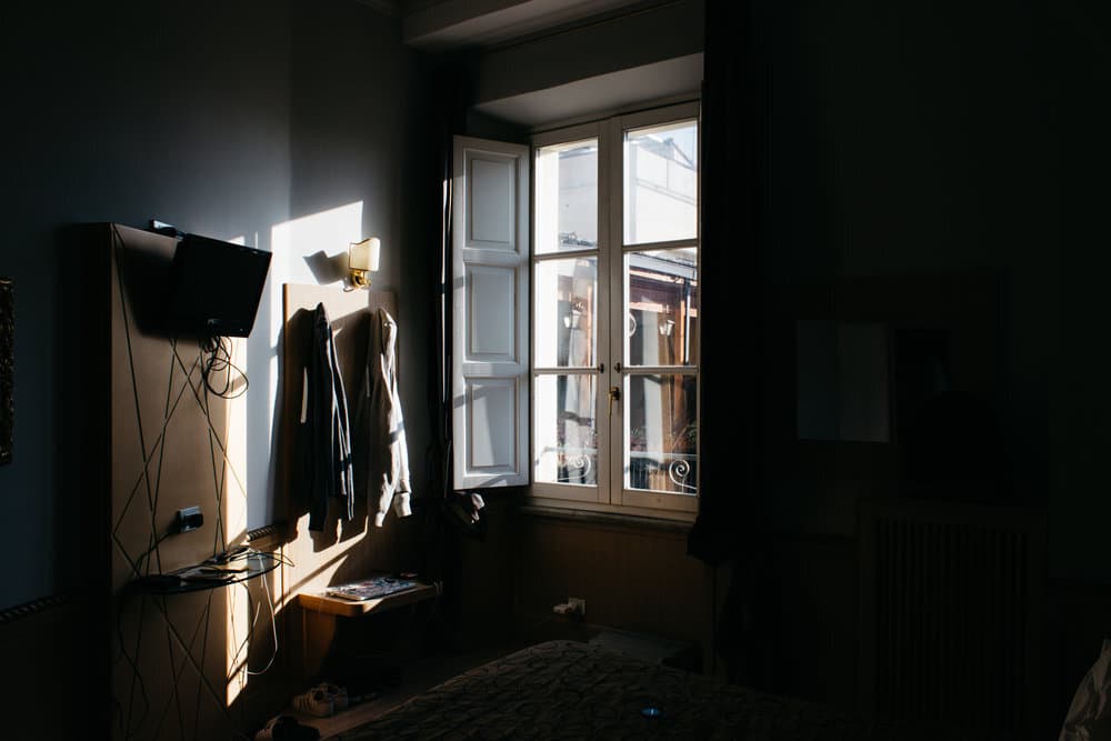 A dimly lit room with an open window, sunlight casting shadows on the wall, and a mounted TV.