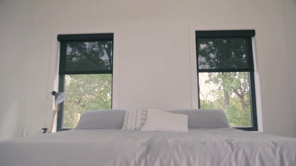 Minimalist bedroom with a neatly made bed and two large windows featuring black roller shades, offering a view of trees outside.