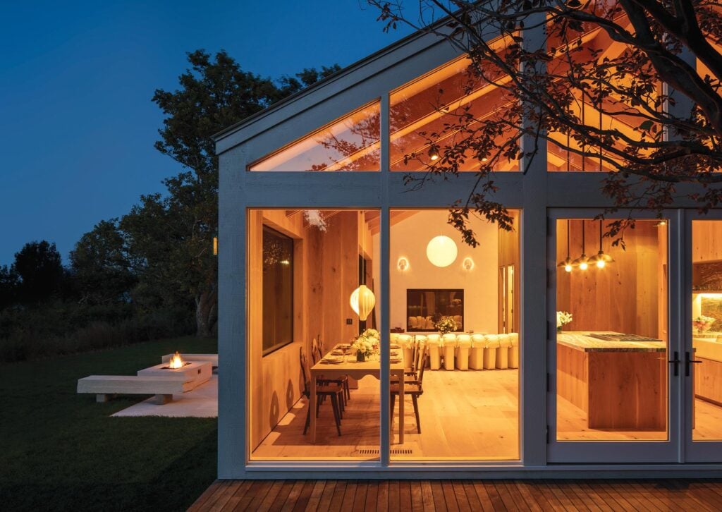 Modern home with large glass windows glowing warmly at dusk, showcasing a cozy dining area, living room, and kitchen with elegant lighting and wooden interior accents.