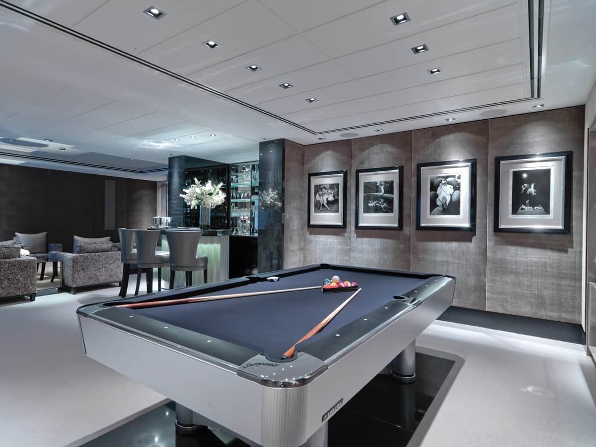 A modern luxury game room featuring a sleek black and silver pool table in the center, with pool cues resting on it. Behind the table is a stylish bar area with a glowing counter and floral centerpiece, flanked by a mirrored glass cabinet stocked with bottles and glassware. The back wall is decorated with framed black-and-white sports photography, and there are cozy seating areas with patterned chairs to the side. The ceiling is fitted with recessed lighting, adding to the elegant ambiance.