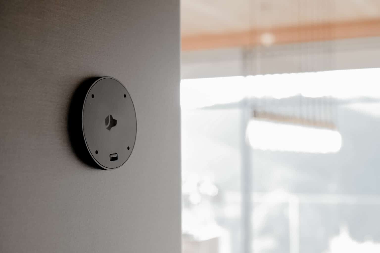 A close-up view of a black round smart device mount attached to a gray wall, with a USB-C port at the bottom. The background features a blurred, modern, sunlit interior with large windows and contemporary lighting.