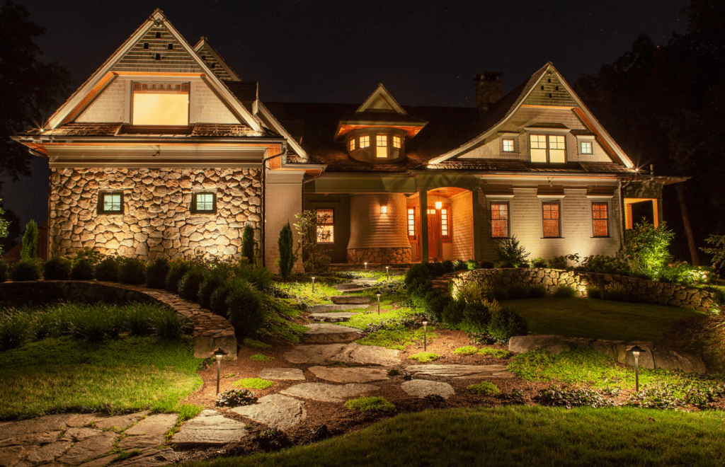 A charming, stone-accented cottage-style home beautifully illuminated at night. Warm outdoor lighting highlights the architectural details, pathway, and landscaping, creating a welcoming ambiance. The house features gabled roofs, wooden trim, and glowing windows that give off a cozy and elegant atmosphere. A stone walkway leads to the front porch surrounded by manicured garden beds and low landscape lighting.