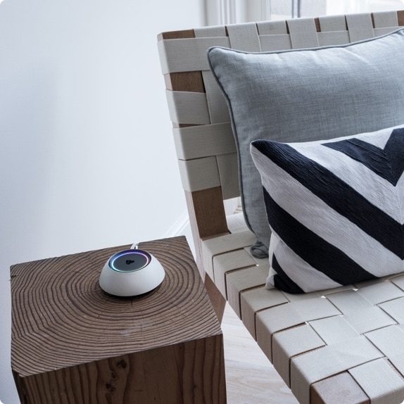 A small modern smart home device with a glowing ring sits on a wooden side table next to a woven accent chair. The chair has a light cushion and a bold black-and-white chevron pillow, adding a touch of contemporary design to the minimalist room.
