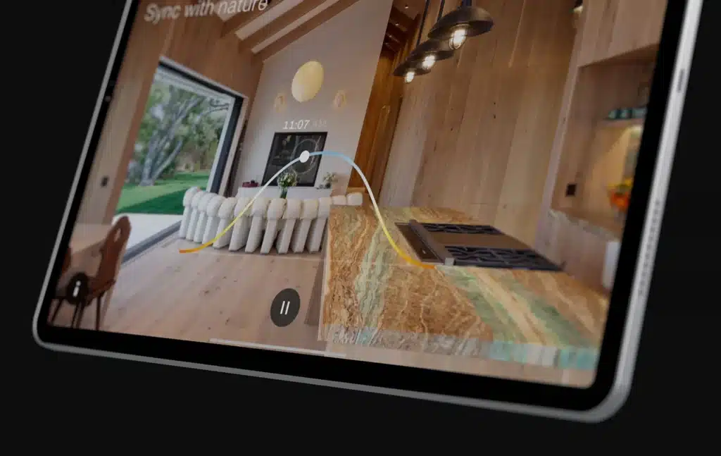 Close-up of a tablet displaying a home automation interface with a daylight curve graph overlayed on a modern kitchen and living area. The app shows natural lighting sync controls with a timeline set to 11:07 AM. The room features wooden finishes, a stone kitchen island, and large open windows overlooking a green yard.