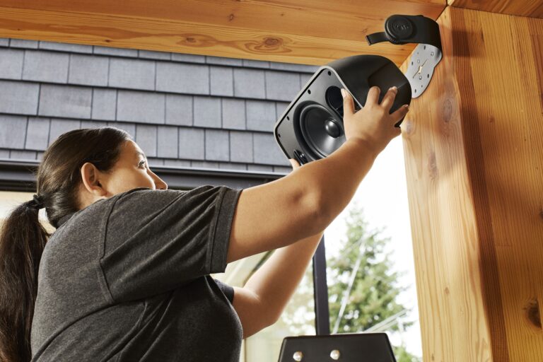 A woman in a gray shirt installs a large black speaker onto a wooden outdoor wall using a mounted bracket. She is reaching up with both hands to secure the speaker while standing on a ladder. The background shows a window, shingles on a house exterior, and trees outside.