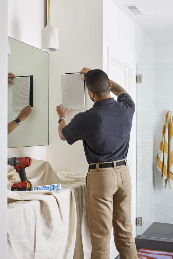 A man installs a smart wall panel or speaker in a modern bathroom, with tools on a covered countertop and a reflection visible in the mirror.