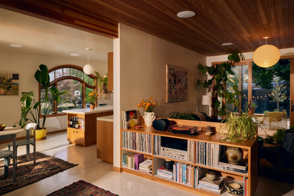 A cozy, plant-filled open-concept home interior featuring a kitchen, dining, and living area. The kitchen has warm wood cabinets and modern lighting. A large arched window brings in natural light. The dining table is surrounded by chairs and indoor plants. The living area includes a bookshelf filled with vinyl records and books, with a record player and a wireless speaker on top. A large sliding glass door leads to a patio lit with string lights. Ceiling-mounted speakers are visible, indicating an integrated sound system.