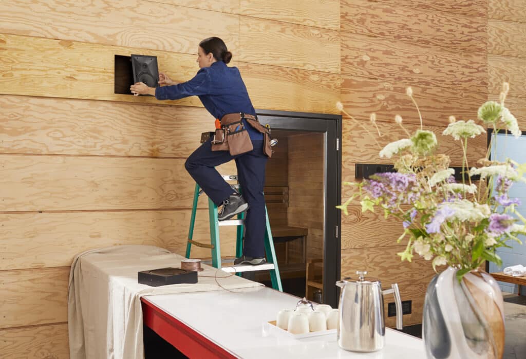 A woman on a ladder installing a speaker into a wooden wall inside a modern home, with tools around and flowers in the foreground.