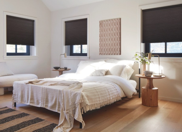 Cozy bedroom with three windows featuring dark cellular shades and a neatly made bed with neutral bedding.