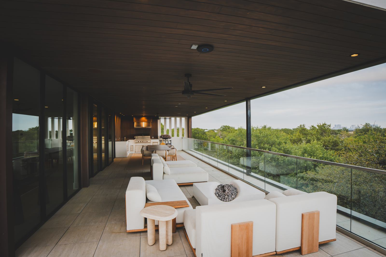 Covered upper-level patio with white outdoor sofas, ceiling fans and a glass rail overlooking trees